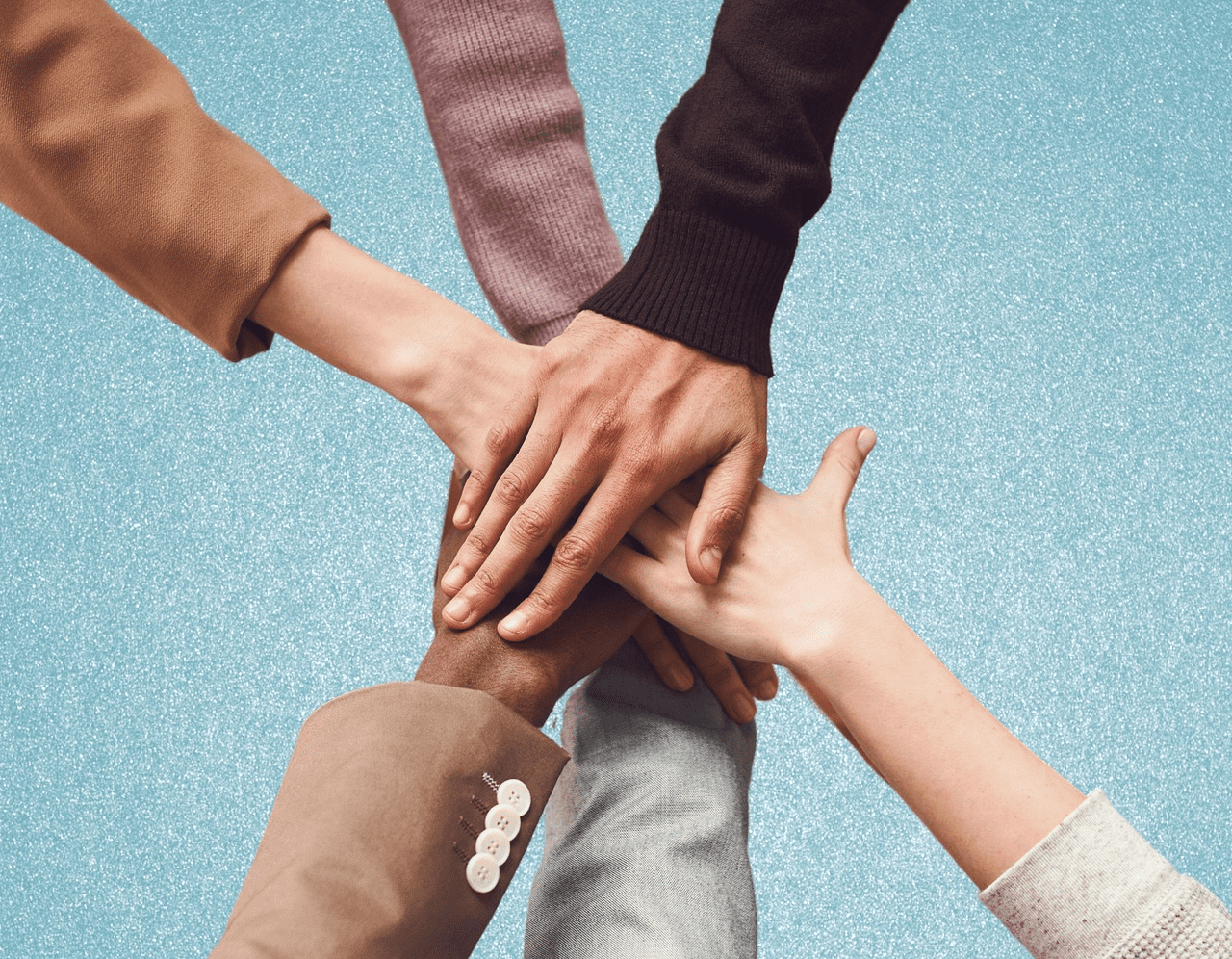 non profit 1 A diverse group of six hands, belonging to people of different skin tones and genders, join together in a supportive gesture against a light blue background. Each person's arm is dressed differently, highlighting a mix of styles and colors.