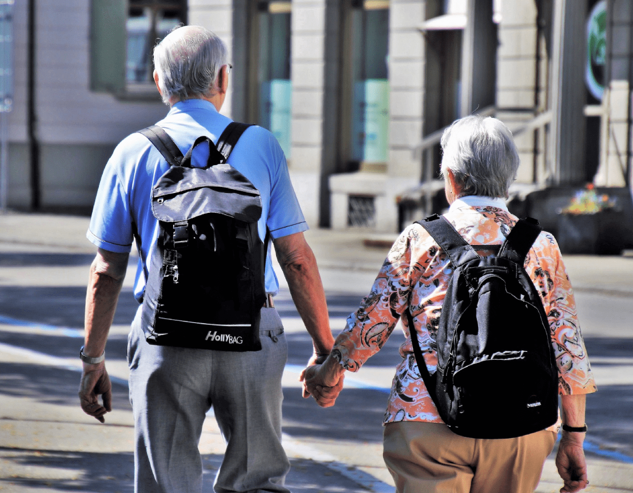 non profit 23 An elderly couple walking hand in hand on a sunny sidewalk. Both have gray hair and are wearing backpacks. The man is wearing a blue polo shirt and gray pants, and the woman is wearing a floral blouse and beige pants. They are seen from behind.