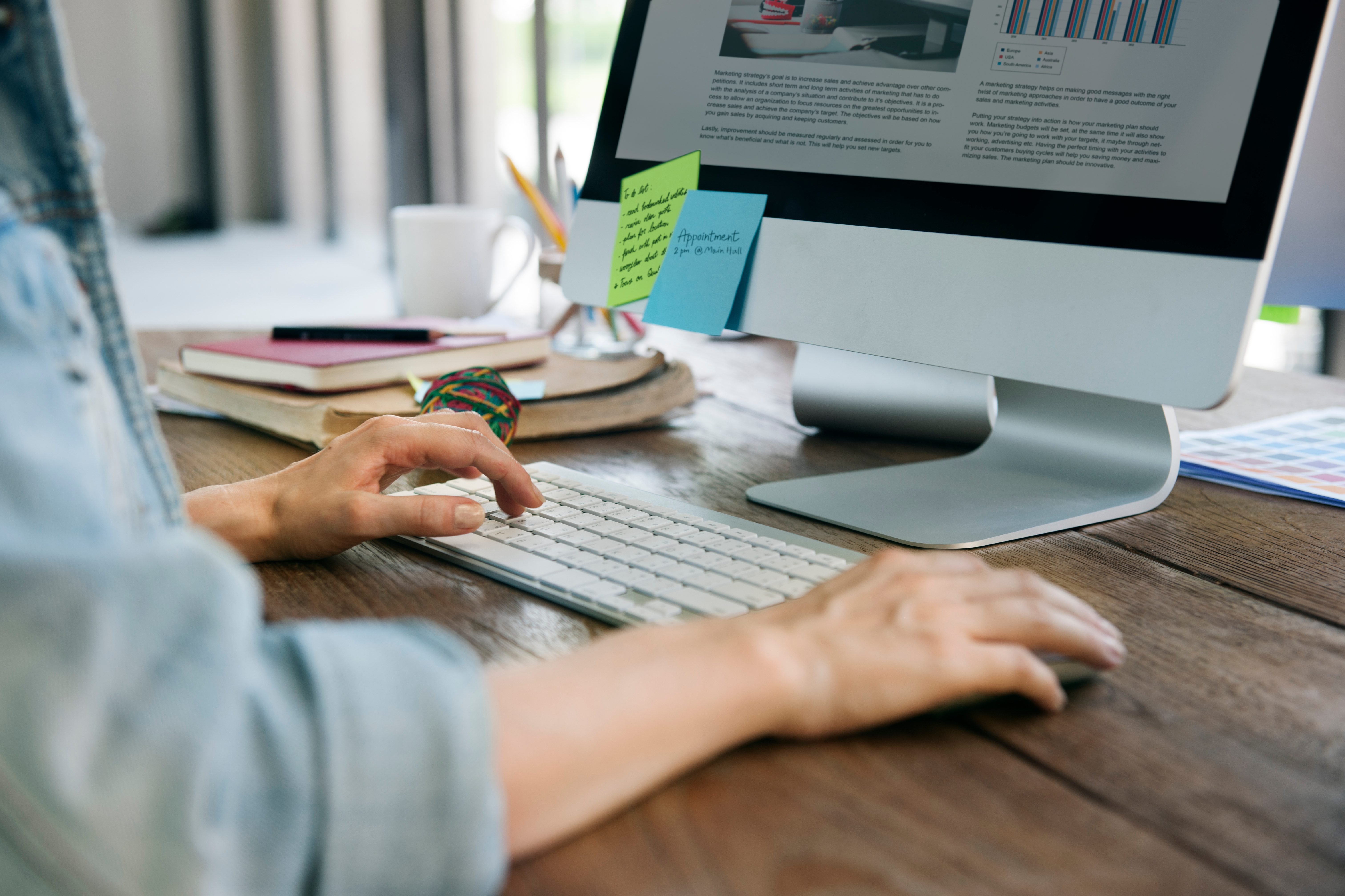 A person seated at a desk with dual monitors displaying text and coding, surrounded by tech equipment and a plant.